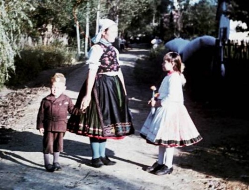 Egy asszony l�that� a k�pen, s�t�t, d�szes n�pviseletben. Egy kisfi� kez�t fogja, akinek ruh�zata egyszer�, barna kab�t �s t�rdnadr�g. Az asszonnyal szemben egy 10-12 �ves l�ny l�that�. Ruh�zata vil�gos, feh�r, minta csak a szoknya perem�n l�that�.