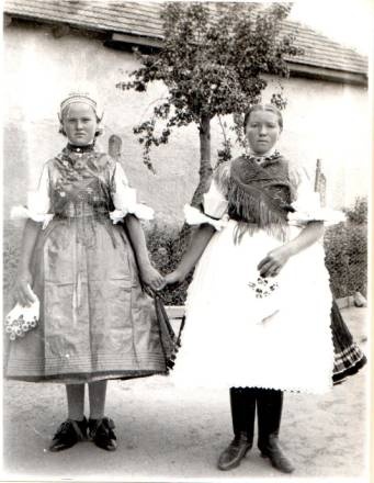 Két népviseletbe öltözött nő áll egymás mellett. Egymás kezét fogva. Régi, fekete-fehér felvétel.