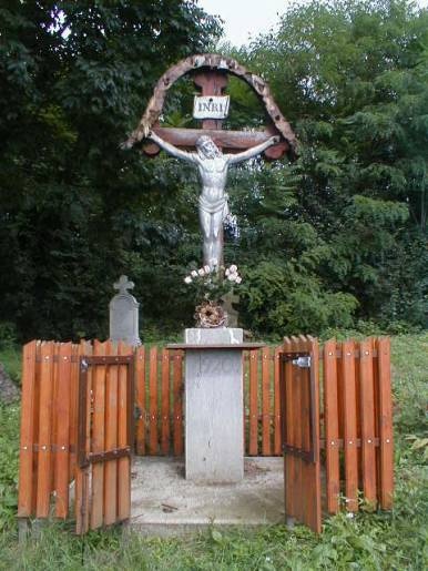A megfeszített Jézust ábrázoló, kőoszlopon álló feszület. Körben világosbarna léckerítés, nyitott kapuval.