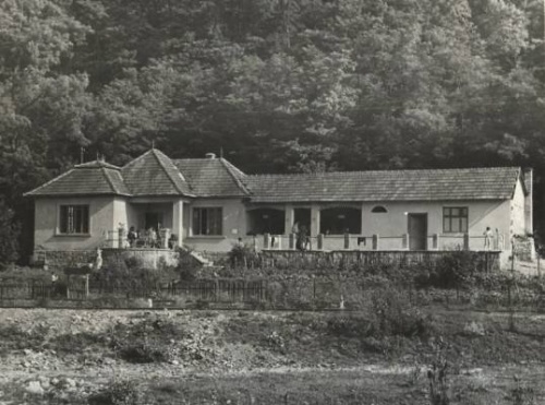 Hosszk�s, feh�rre meszelt, torn�cos h�z terasszal. A h�tt�rben domboldal, erd�vel.