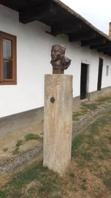 A Bánki menyecske szobra. Női fejet ábrázoló szobor kőoszlopon.